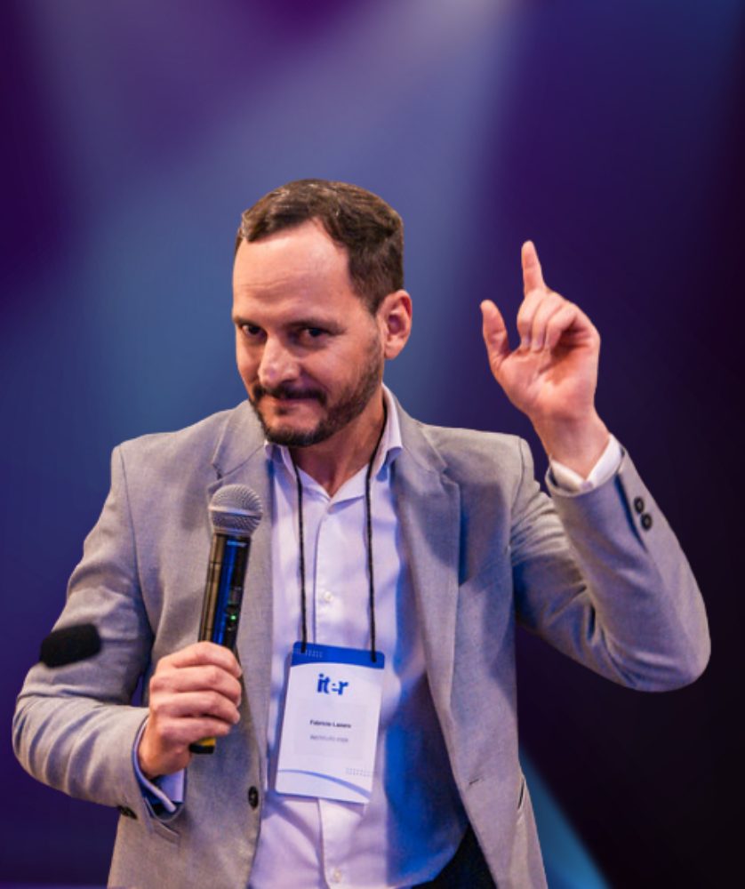 Fabrício Lázaro falando ao microfone em um palco com luzes roxas